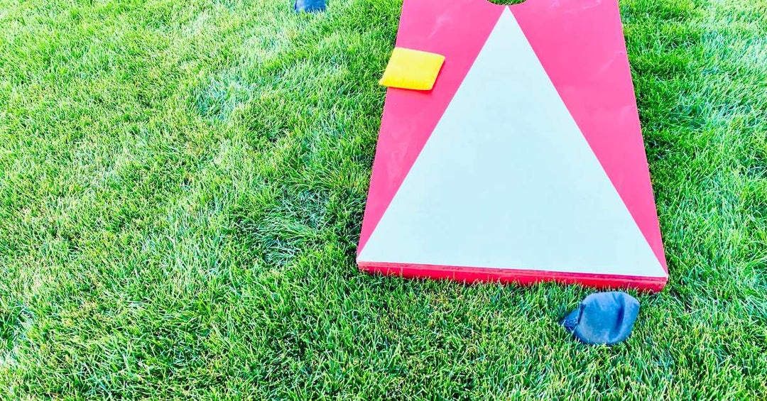 A red and white painted cornhole board sits on the grass. There are blue and yellow cornhole bags strewn around it.