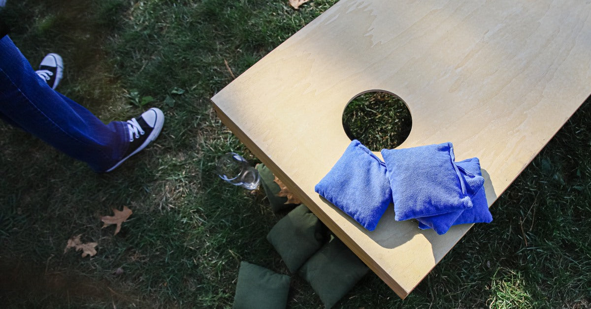 An overhead view of a cornhole board with blue bags on it. There are green bags on the grass and someone standing next to the board.