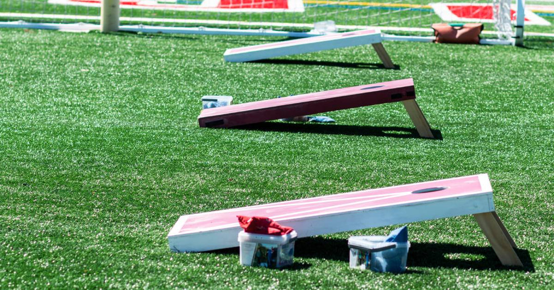 Three cornhole boards lined up next to each other on a grassy field. They have sets of bags sitting next to them.