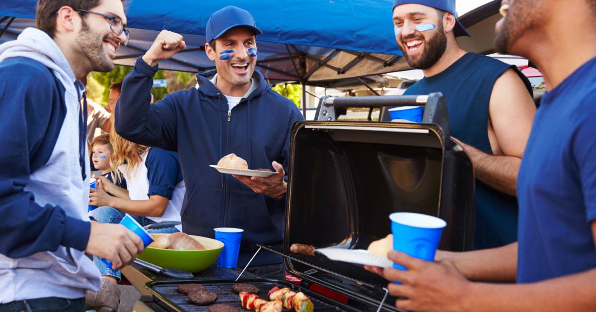 A group of guys surrounding a grill at a tailgating party. They all have either food or cups in their hands.