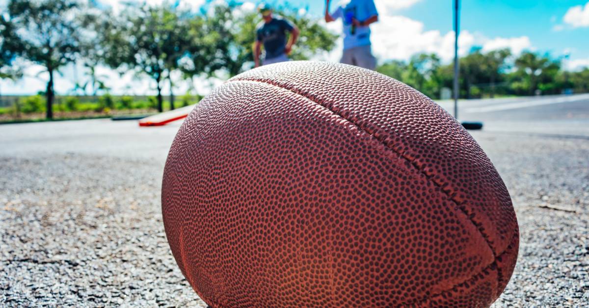 A close-up shot of a football sitting on asphalt. There are two men playing cornhole in the background.