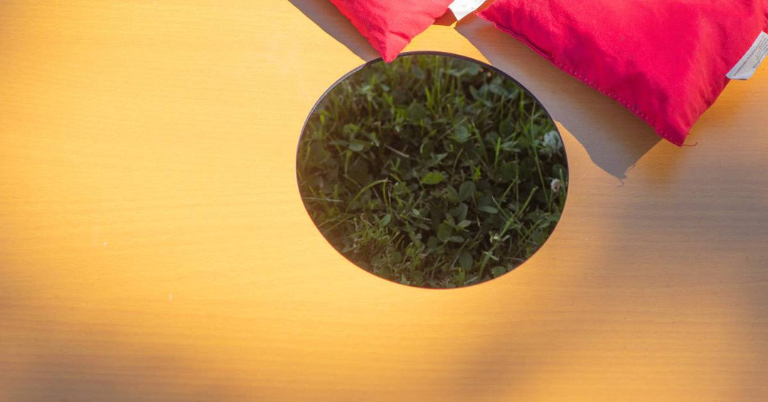 A close-up of a yellow cornhole board hole. The hole is positioned over grass with some red bags around it.