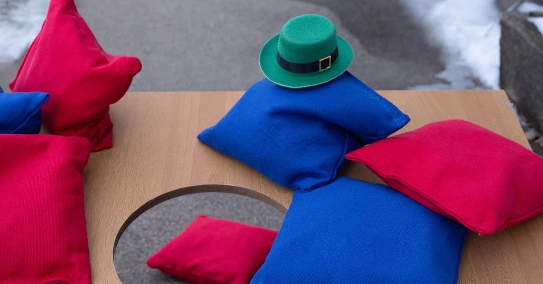 Red and blue cornhole bags sitting on top of a board. One of the blue bags has a little green hat sitting on it.