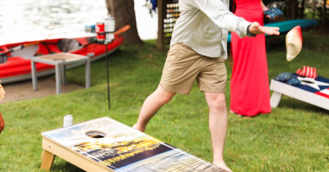 Side-by-side cornhole boards with people playing at them during a tournament. There is a lake in the background.